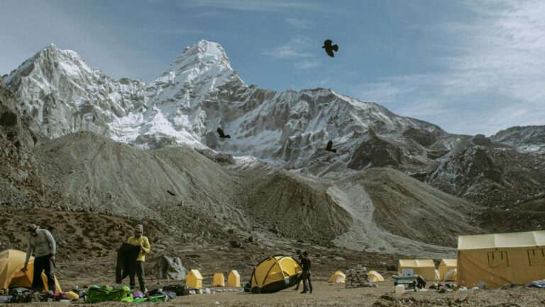 Campers preparing tents at a scenic mountain campsite with snowy peaks and birds flying.