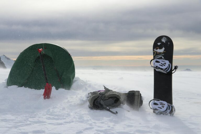 Snowy mountain camping setup with a tent, snowboard, and backpack in winter.