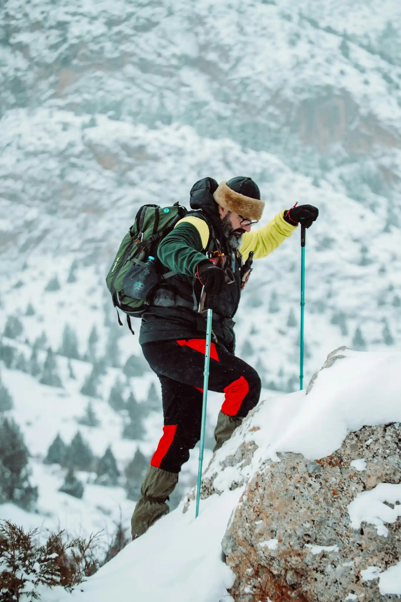 Male mountaineer in snow gear climbing a rocky slope with trekking poles.